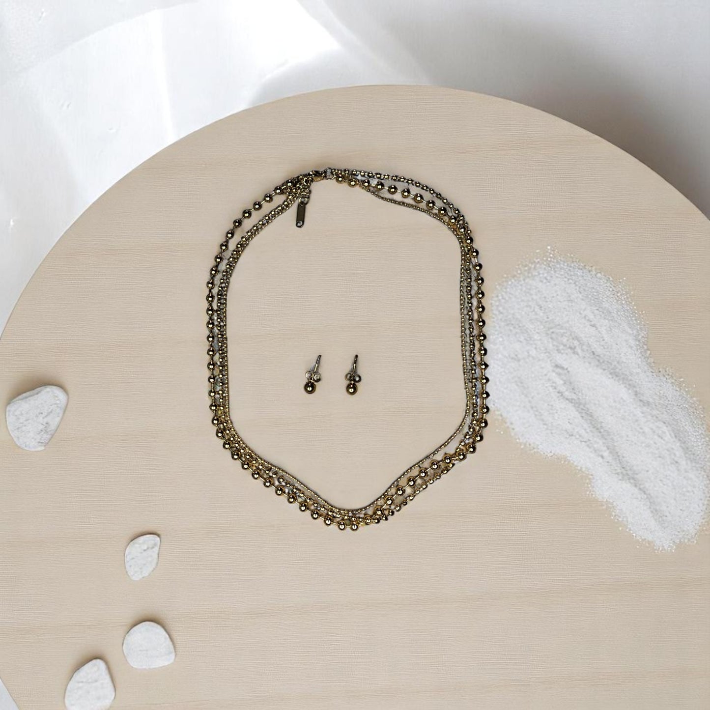 Joyería dorada con diseño de balines y aretes tipo topo sobre fondo de arena
