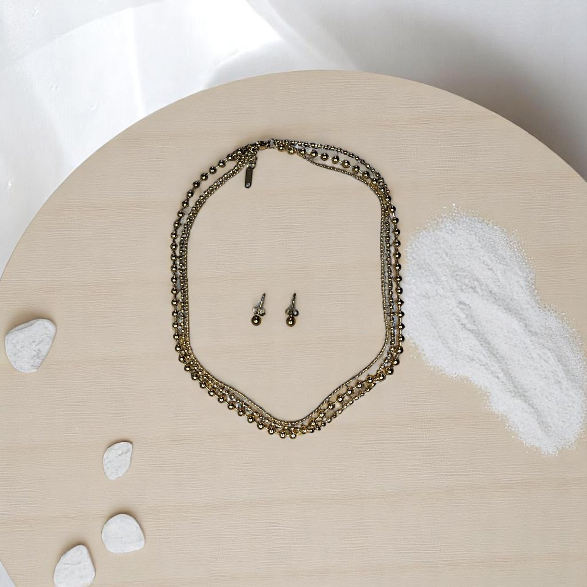 Joyería dorada con diseño de balines y aretes tipo topo sobre fondo de arena